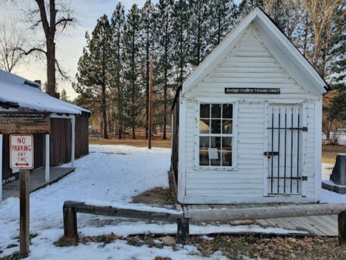A small white historic house with a sign reading "Judge Halley House 1867," surrounded by snow and trees.