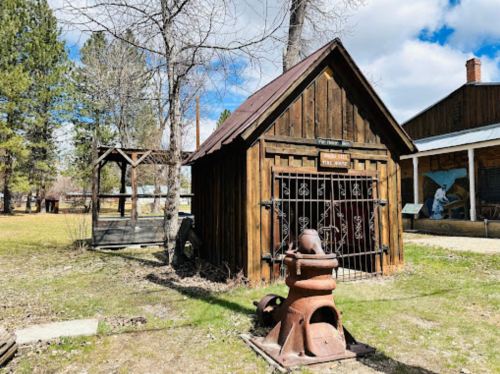 A small wooden building with a metal gate, surrounded by trees and grass, featuring an old forge in the foreground.