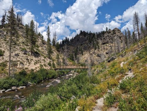 A wooden bridge spans a rocky stream, surrounded by lush greenery and mountains under a partly cloudy sky.