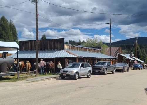 A rustic street scene with parked cars and people near wooden buildings, with horses in the background under a cloudy sky.
