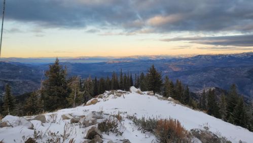 A scenic mountain view at sunset, featuring snow-covered ground and distant peaks under a cloudy sky.