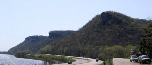 Scenic view of a winding road alongside a river, with lush green hills and rocky cliffs in the background.