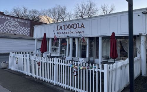 Exterior of La Tavola restaurant with outdoor seating, red umbrellas, and a white picket fence. Open sign visible.