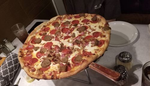A large pizza topped with pepperoni, sausage, and cheese, served on a wooden pizza stand in a restaurant setting.