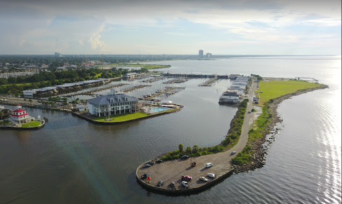 Aerial view of a marina with boats, green land, and a waterfront road under a cloudy sky.