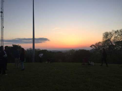 Silhouetted figures on a grassy hill at sunset, with a colorful sky and distant trees in the background.