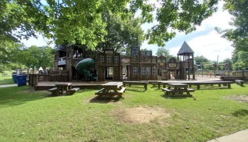 A wooden playground with slides and play structures, surrounded by green grass and picnic tables under leafy trees.