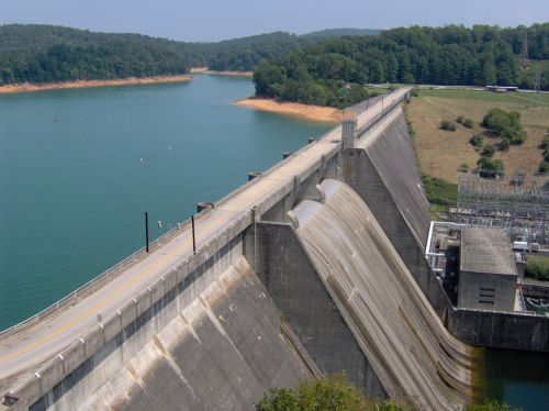 Aerial view of a concrete dam with water flowing over it, surrounded by a lake and green hills in the background.