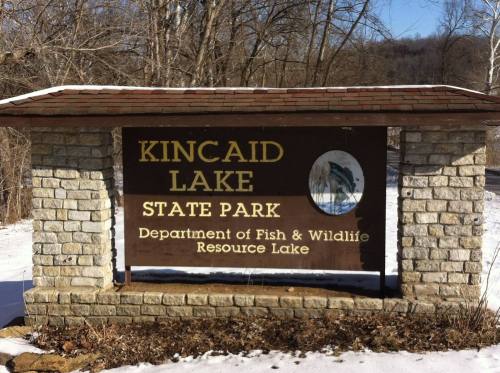Sign for Kincaid Lake State Park, featuring a stone structure and snowy surroundings, with a fish and wildlife department logo.