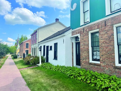 A charming street lined with historic brick and white houses, surrounded by green grass and blue skies.
