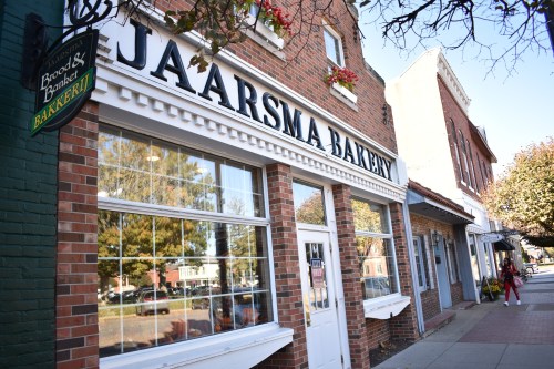 A brick bakery storefront with large windows and a sign reading "Jaarsma Bakery," surrounded by trees and shops.