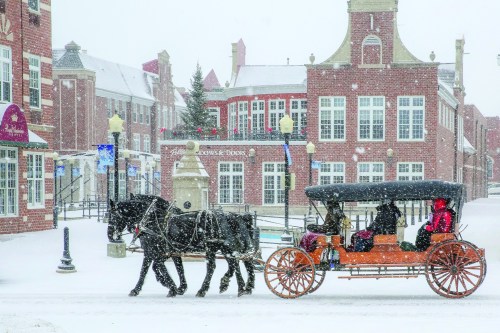 A horse-drawn carriage travels through a snowy street, with historic buildings in the background. Snowflakes are falling.