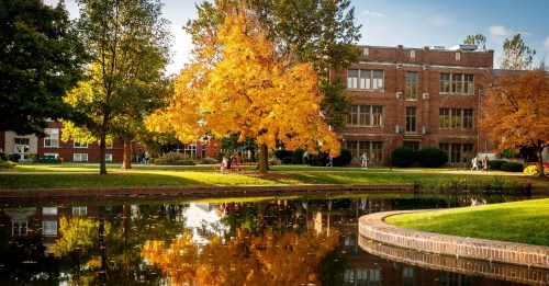 A serene campus scene with a vibrant autumn tree reflected in a pond, surrounded by brick buildings and students.