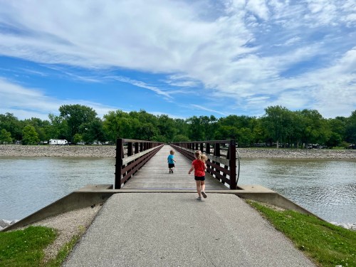 Two children walk on a wooden bridge over a calm river, surrounded by greenery and a blue sky with clouds.