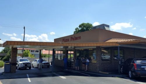 Exterior of Pizza Palace restaurant with customers at the counter and cars parked outside under a clear blue sky.