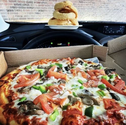 A pizza with vegetables sits in a car, with a plate of bagels in the background on the dashboard.