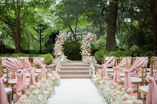 A beautifully decorated outdoor wedding aisle with floral arrangements and elegant chairs adorned with pink ribbons.