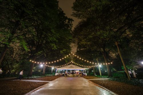 A beautifully lit outdoor event space with string lights, surrounded by trees at night. Guests gather under a tent.