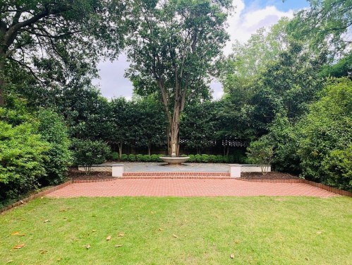 A serene garden space with a central fountain, surrounded by lush greenery and a brick patio.