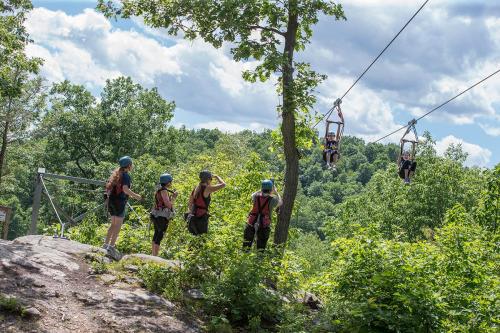 A group of people in helmets watches two individuals ziplining through a lush green landscape under a cloudy sky.