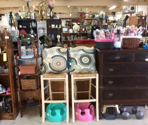 A cluttered thrift store display featuring furniture, decorative items, and colorful cushions on bar stools.