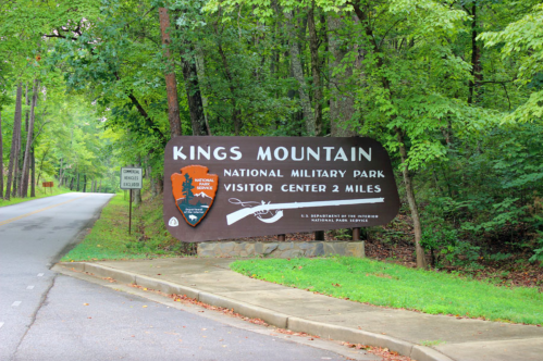 Sign for Kings Mountain National Military Park, indicating the visitor center is 2 miles ahead, surrounded by trees.