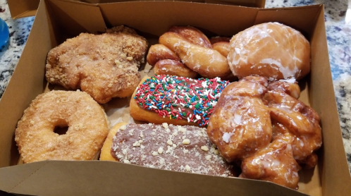 A box of assorted donuts, including glazed, sprinkled, and cinnamon varieties, arranged on a countertop.