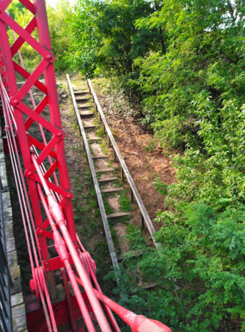 A red metal structure overlooks a steep, overgrown path with wooden steps leading down through greenery.