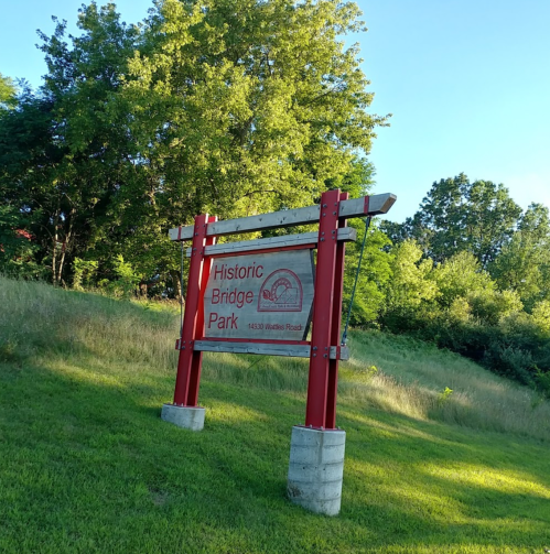 Sign for Historic Bridge Park, featuring red and white colors, surrounded by green grass and trees.