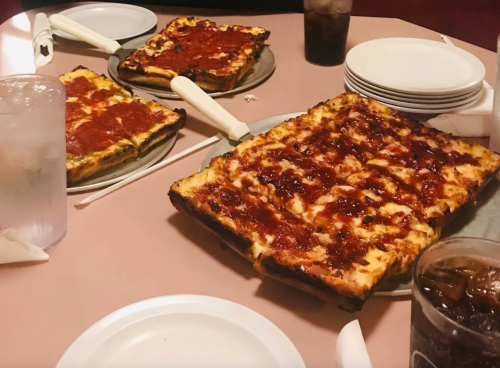 A table with multiple rectangular pizzas topped with sauce, alongside drinks and plates.