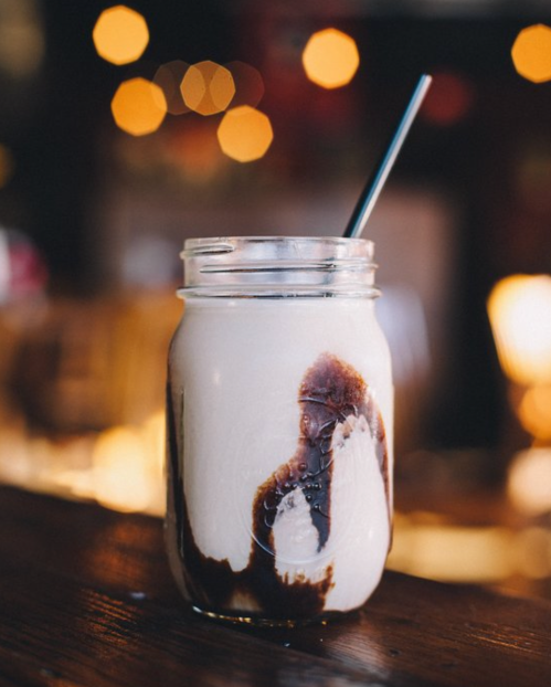 A glass jar filled with a creamy drink, swirled with chocolate syrup, and a straw, set against a blurred background.