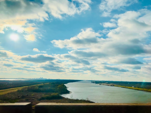 A scenic view of a river winding through a landscape under a bright sky with fluffy clouds and distant city skyline.