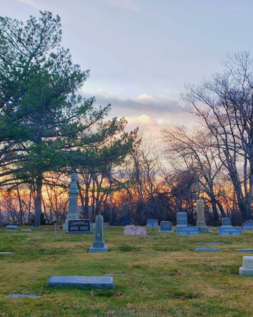 A serene cemetery at sunset, featuring various gravestones and trees against a colorful sky.