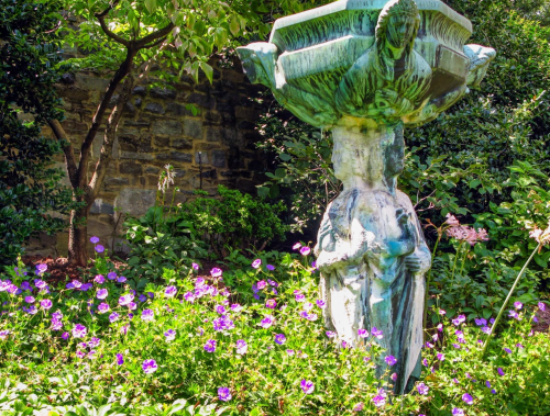 A weathered statue of a woman holding a basin, surrounded by vibrant purple flowers and lush greenery.