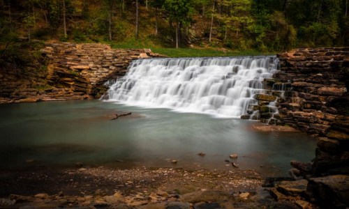 A serene waterfall cascades over rocky ledges into a calm pool, surrounded by lush greenery.