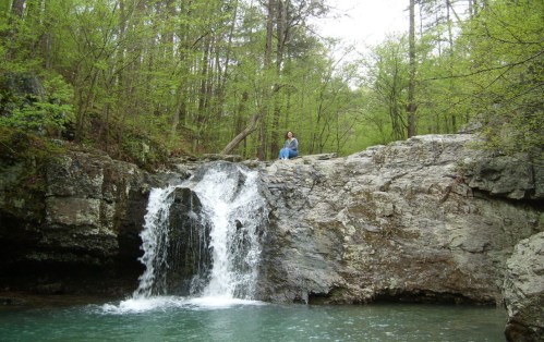 A serene waterfall cascades over rocks into a turquoise pool, surrounded by lush green trees. A person sits nearby.