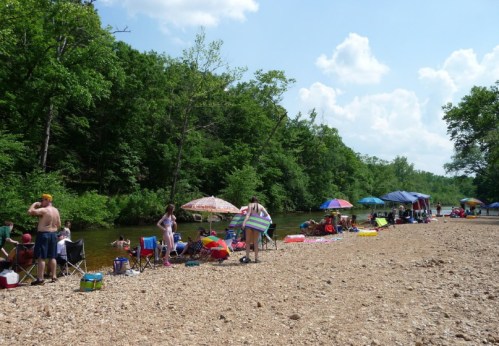People enjoying a sunny day by a river, with umbrellas and chairs set up along the sandy shore.