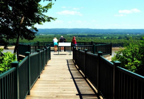 A wooden walkway leads to a scenic overlook with three people enjoying a view of green hills and a blue sky.