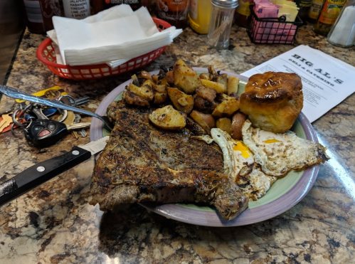 A hearty breakfast plate featuring a large steak, fried egg, seasoned potatoes, and a biscuit on a colorful plate.