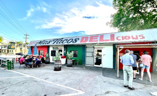 A colorful taco deli with outdoor seating, surrounded by greenery and barrels, bustling with customers.