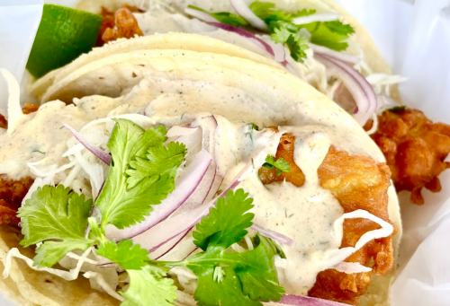 Two tacos filled with crispy fish, topped with creamy sauce, cilantro, red onion, and lime wedges.