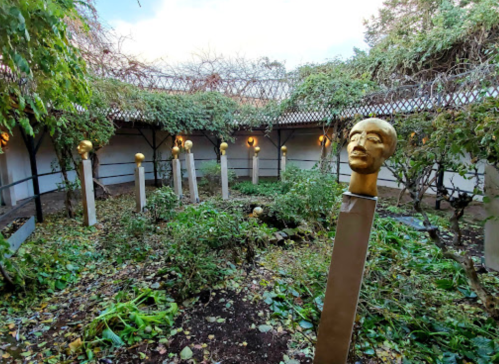 A garden with overgrown plants and several golden busts on tall pedestals amidst a serene, enclosed space.