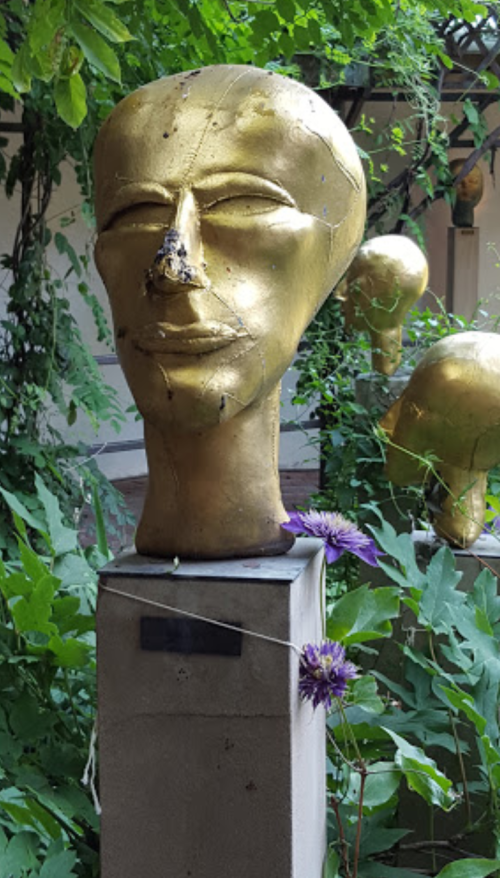 A golden sculpture of a head on a pedestal, surrounded by greenery and a purple flower.