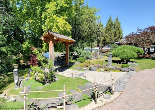 A serene garden featuring a wooden arch, lush greenery, rocks, and a winding path under a clear blue sky.