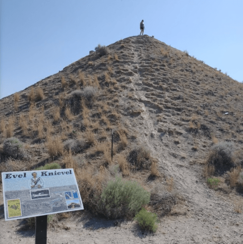 A person stands atop a rocky hill with a sign about Evel Knievel in the foreground, surrounded by dry vegetation.