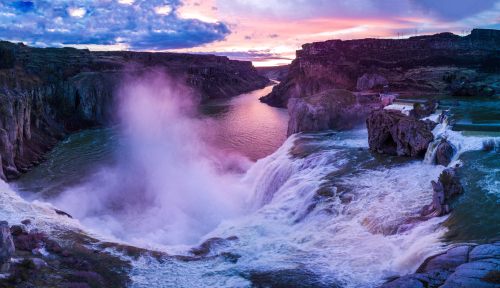 A stunning waterfall cascades into a river at sunset, surrounded by rocky cliffs and a colorful sky.