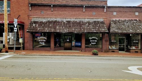 A brick storefront with a shingled roof, featuring "Open" and "Pizza" signs, and a flower pot by the entrance.