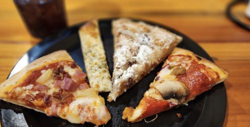 A black plate with five slices of pizza, including toppings like ham, mushrooms, and a dessert slice with powdered sugar.