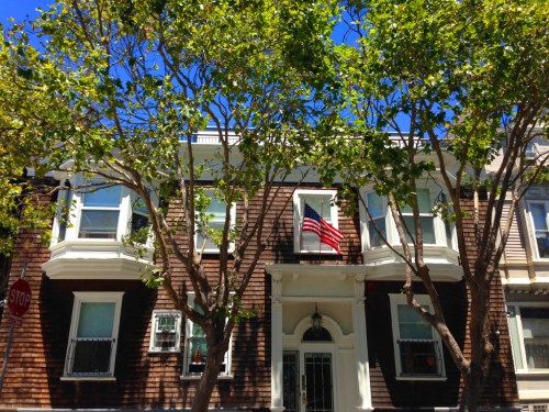 A brownstone building with large trees in front, featuring an American flag hanging from a window under a clear blue sky.