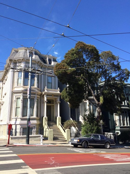 Victorian-style house with a large tree, blue sky, and power lines, located at a street intersection.